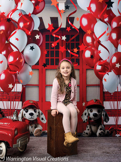 Happy girl sitting front and center with red and white balloons and firehouse backdrop featuring stuffed dalmatians and red fire station doors.
