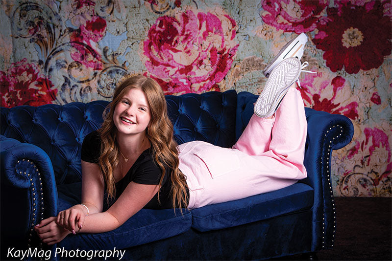 Teen girl lounging on a navy blue tufted velvet couch in front of a large floral-patterned backdrop, ideal for fashion-forward portrait photography.