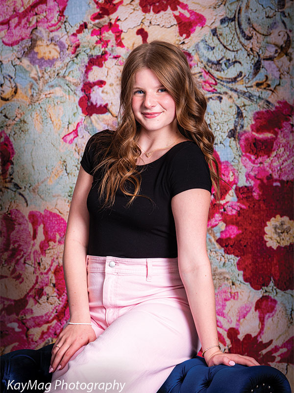 Teen girl standing confidently in front of a vibrant floral tapestry backdrop with distressed texture, wearing pink pants and a black top—perfect for colorful, bold senior portraits.