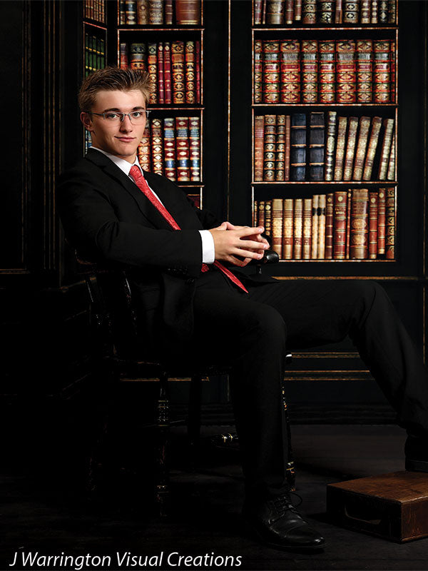 Teen boy seated with hands folded in a studio setting using a formal library-themed backdrop, ideal for traditional senior or formal portraits.