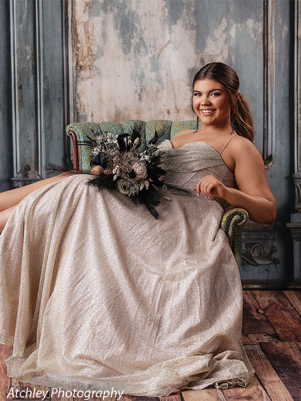 Hand-painted French panel backdrop with antique textures, featuring a female senior in a gold gown holding a bouquet, seated in a blue-green armchair for formal portraits.
