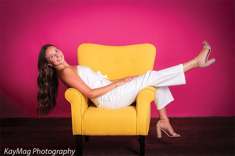 Stylish young woman reclining in a yellow accent chair with a vivid fuchsia pink backdrop, perfect for modern senior portraits or editorial-style photo shoots.