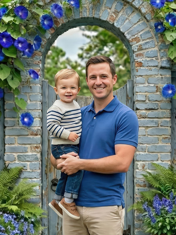 Man wearing a blue polo shirt holds a smiling toddler dressed in a striped sweater while standing beneath a brick garden archway backdrop decorated with blue flowers, greenery, and a painted wooden door.