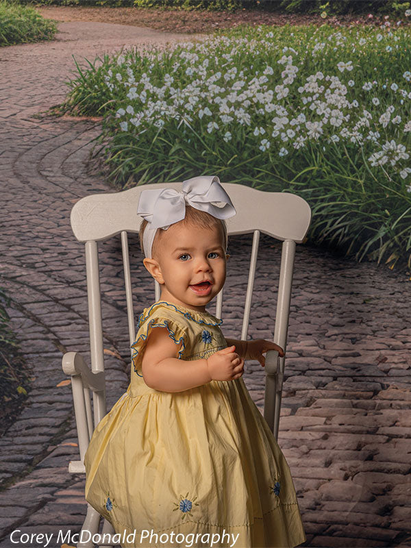 Baby girl wearing a yellow dress with blue floral embroidery and a large white bow headband stands beside a white wooden chair on a curved brick garden path backdrop lined with white flowers and green grass.