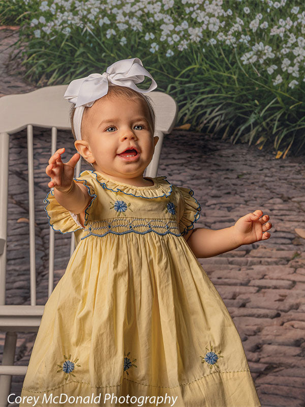 Baby girl wearing a yellow dress with blue floral embroidery and a white bow headband reaches forward while standing near a white wooden chair on a brick garden walkway backdrop lined with white flowers and greenery.