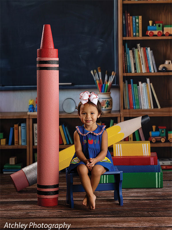 Giant red crayon and yellow pencil photography props in a back-to-school classroom set, the crayon standing upright beside a toddler girl seated on a small blue bench GCP