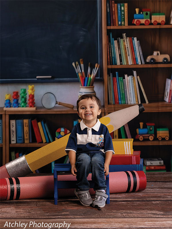 Giant red crayon and yellow pencil photography props framing a laughing toddler boy on a small blue bench, the crayon lying on the wood floor in front of him GCP