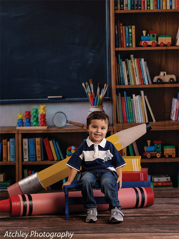 Giant red crayon and yellow pencil photography props arranged in a classroom set, the crayon lying in front of a small bench where a smiling toddler boy sits GCP