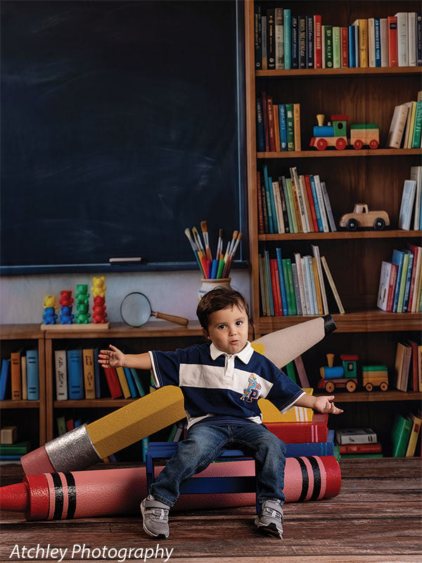 Giant red crayon lying on the floor and yellow pencil propped on oversized books, used as photography props with a toddler boy seated on a bench with arms outstretched GCP