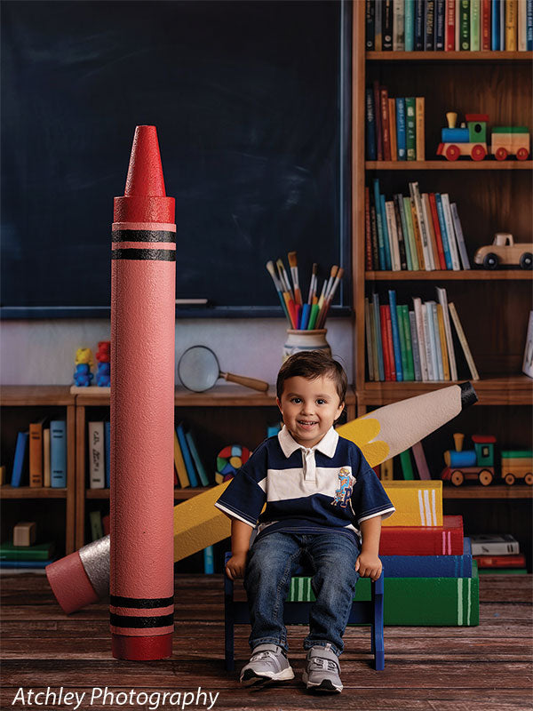 Giant red crayon and yellow pencil photography props in a classroom set, the crayon standing upright beside a smiling toddler boy seated on a small blue bench GCP