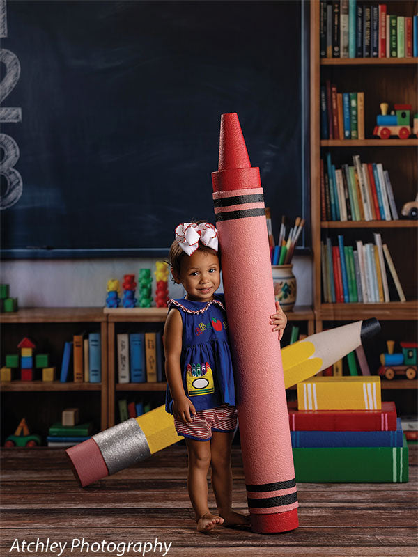 Giant red crayon photography prop standing upright and hugged by a smiling toddler girl, with the giant yellow pencil resting on oversized books in a classroom set GCP