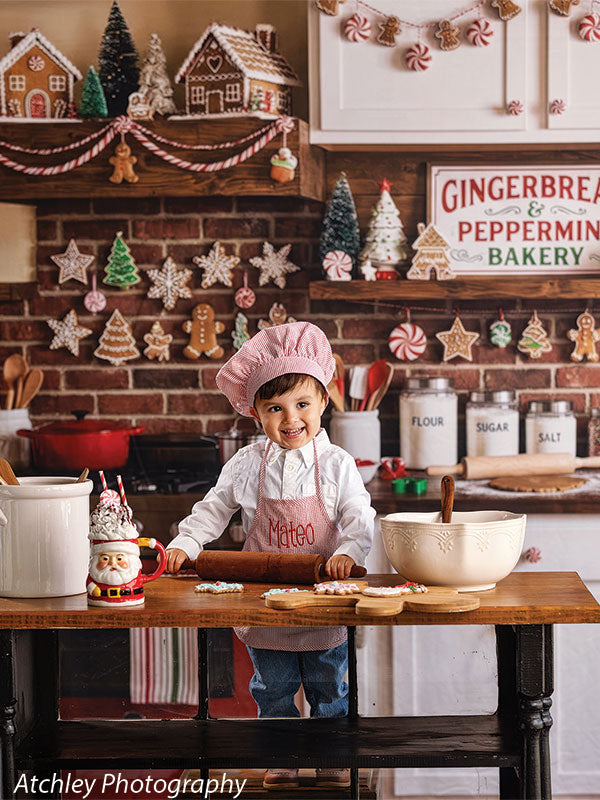 Ginger Street gingerbread bakery kitchen photography backdrop shown with a young boy in a pink chef hat and apron smiling at the camera while baking CP-8876