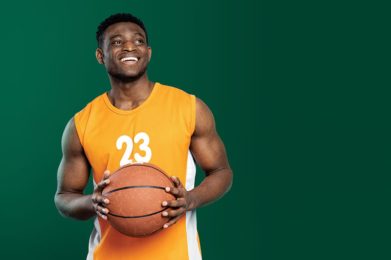 A basketball player in an orange jersey poses in front of a pine green seamless photography backdrop paper in a studio.