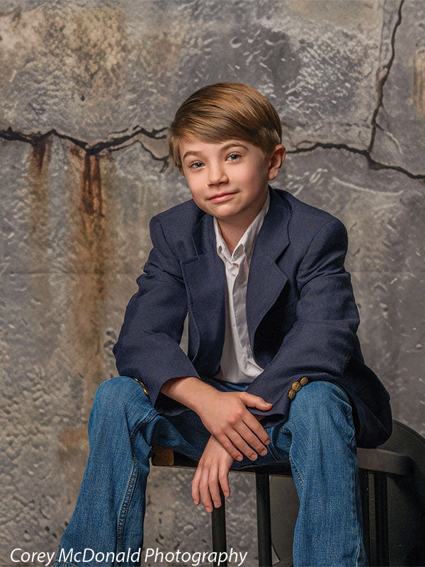Young boy wearing a navy blazer, white shirt, and blue jeans sits on a bench with hands resting together in front of a grunge-style concrete wall backdrop with cracked gray textured surface.