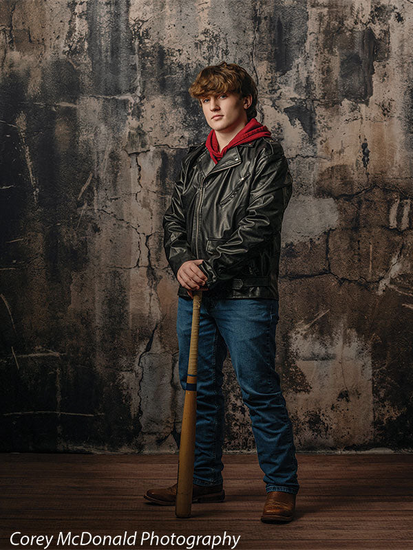 Teen boy with wavy brown hair wearing a black leather jacket over a red hoodie, blue jeans, and brown boots stands holding a wooden baseball bat in front of a dark grunge concrete wall backdrop with cracked industrial texture.