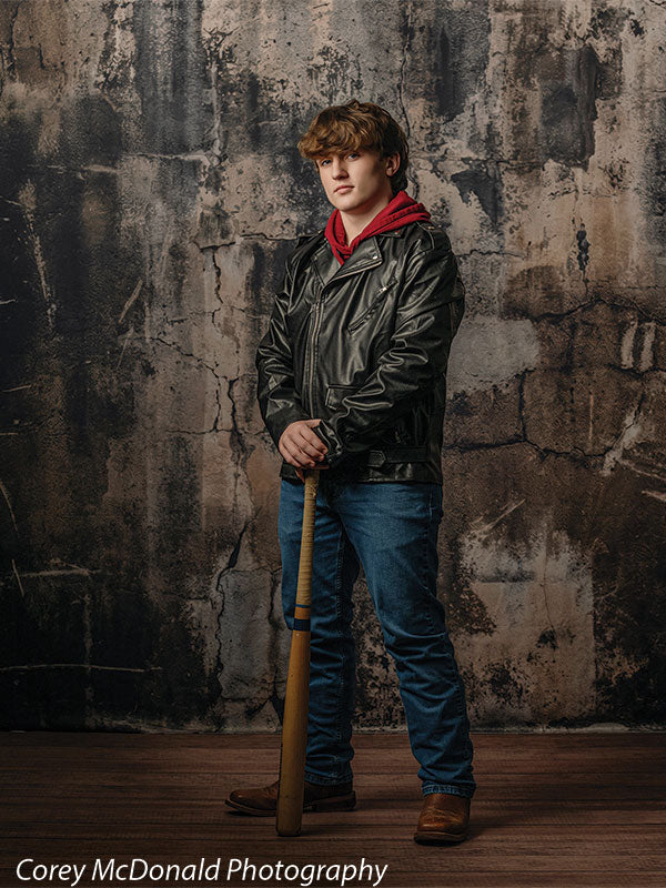 Teen boy wearing a black leather jacket over a red hoodie with blue jeans and brown boots stands holding a wooden baseball bat in front of a dark grunge concrete wall backdrop with heavy texture and industrial tones.