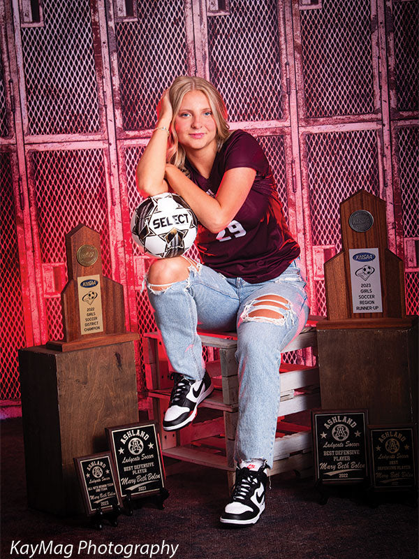 Confident soccer player posing with championship trophies and ball in front of distressed red locker backdrop, ideal for athletic senior sports portraits.