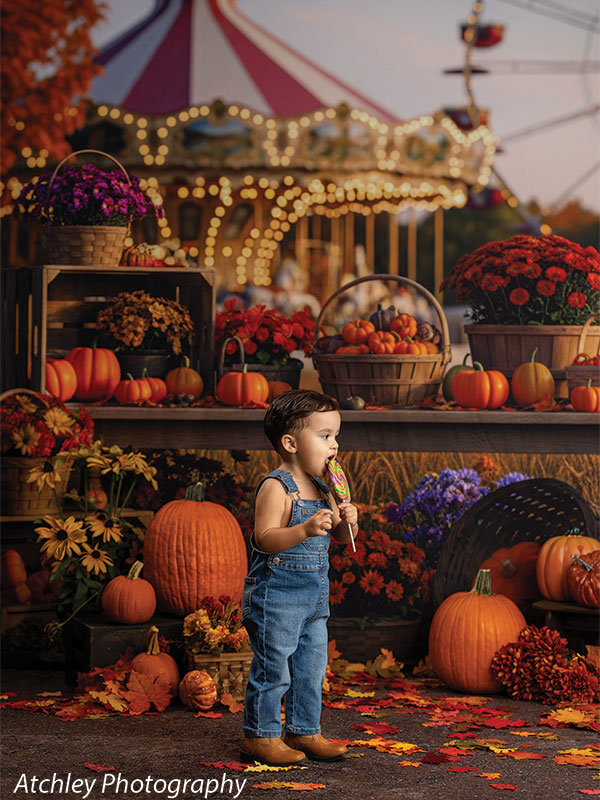 Toddler in denim overalls playing with a wooden toy train on the floor among pumpkins and fall leaves, set before a harvest festival carousel backdrop decorated with baskets of gourds, mums, and warm carnival lights.