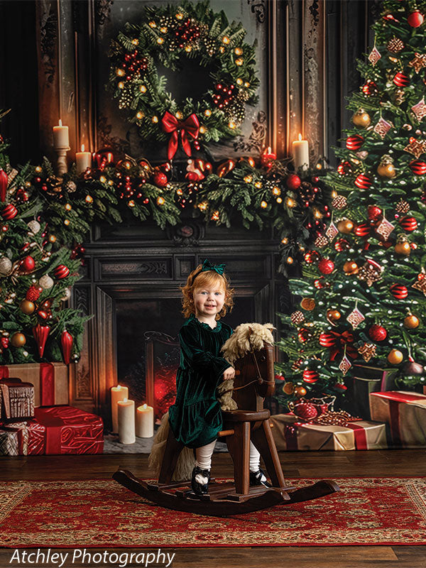 Young girl with curly red hair wearing a dark green velvet dress and white tights sitting on a wooden rocking horse in front of a festive holiday fireplace backdrop featuring a decorated Christmas tree, wreath with red bow, glowing garland, candles, wrapped gifts, and a lit fireplace.