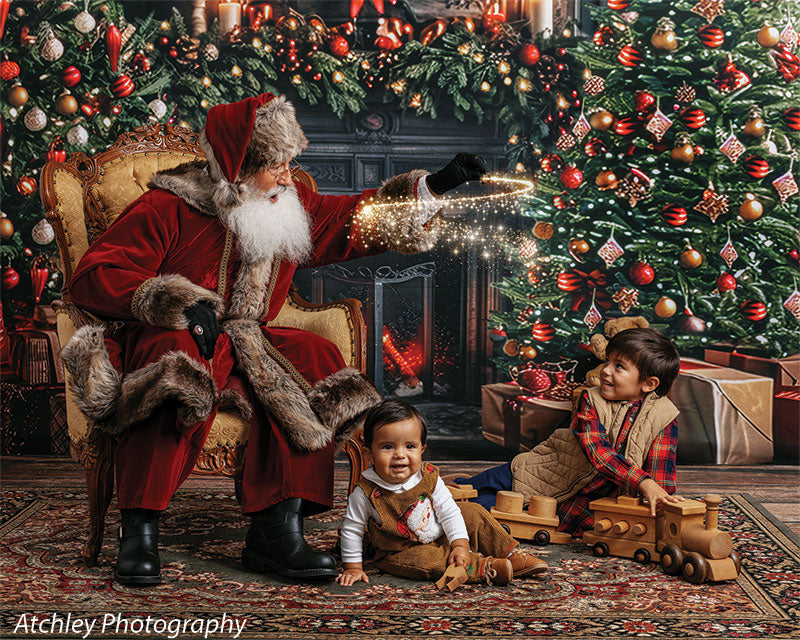 Santa Claus gesturing toward sparkling lights while a baby in brown overalls sits on the floor and a young boy in a plaid shirt and tan vest plays with wooden toy trains, posed in front of a holiday fireplace and Christmas tree backdrop with wreath, garland, candles, and wrapped gifts.