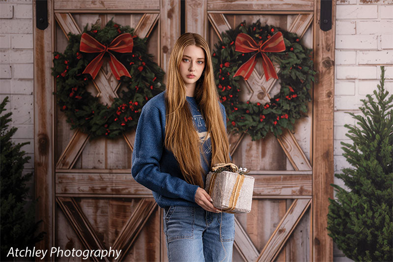 Holly Barn rustic barn doors with Christmas wreaths photography backdrop shown with a senior girl holding a gift in a relaxed pose, wide view CP-8918