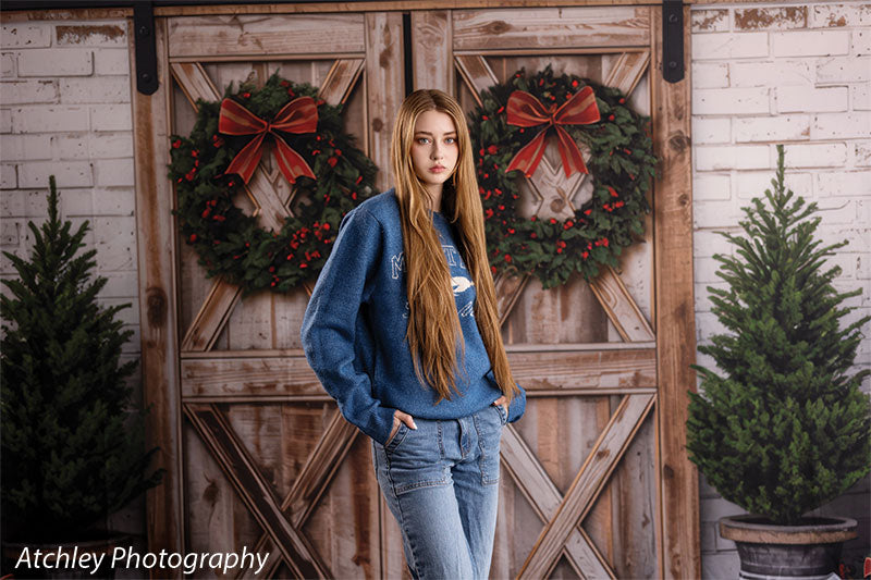 Holly Barn rustic barn doors with Christmas wreaths photography backdrop shown with a senior girl in a blue sweater and jeans, hand in pocket, wide view CP-8918