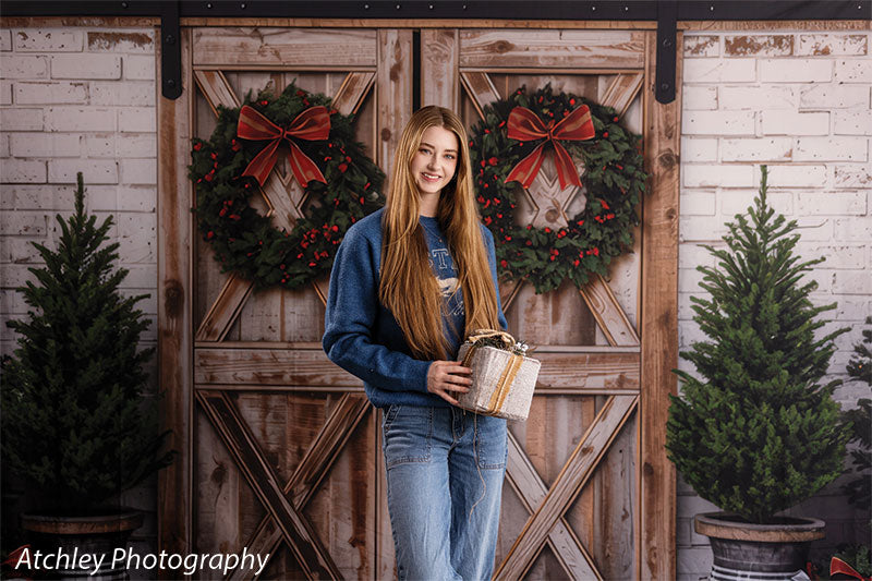 Holly Barn rustic barn doors with Christmas wreaths photography backdrop shown with a senior girl holding a wrapped gift before the wreath-hung doors, wide view CP-8918
