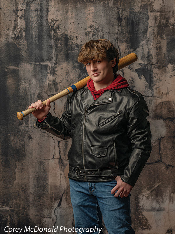 Teen boy in a black leather jacket layered over a red hoodie and blue jeans rests a wooden baseball bat on his shoulder while standing in front of a weathered gray concrete wall backdrop with distressed industrial texture.