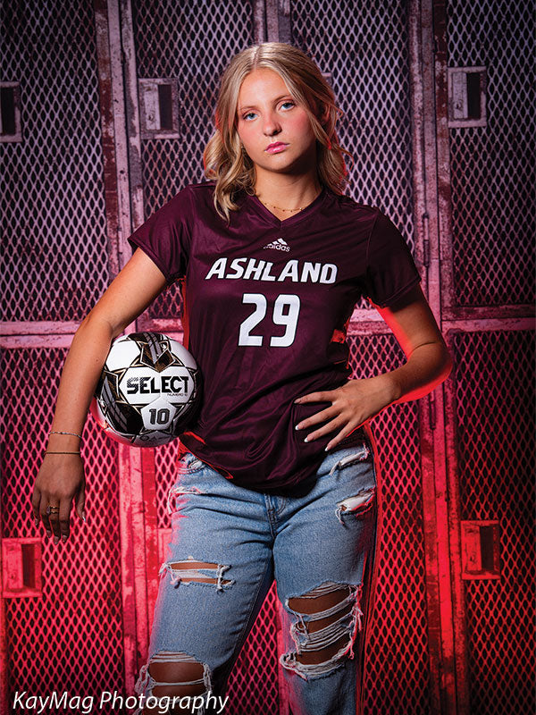 Female soccer player in Ashland jersey posing with ball against red metal locker backdrop, perfect for dynamic sports-themed senior photography.