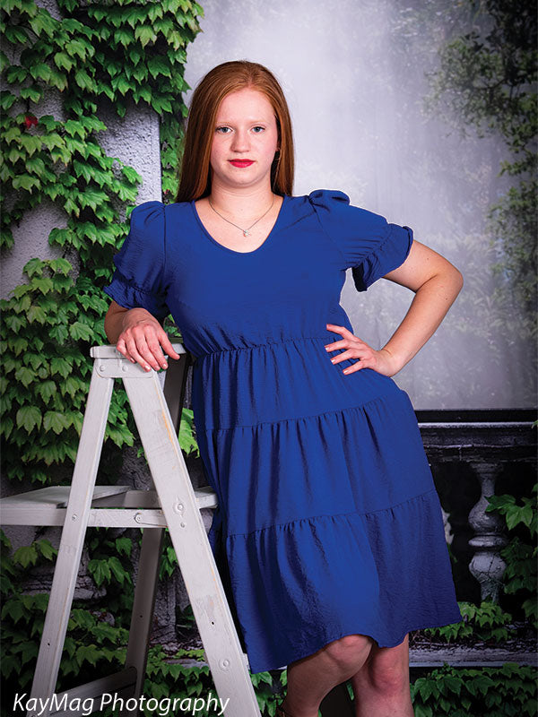 Young woman in a blue dress posing with a white ladder in front of a stone garden wall covered with green ivy, perfect for senior portraits or spring photography sessions.