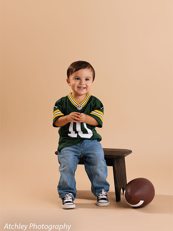 Smiling toddler wearing a green football jersey with the number 10, blue jeans, and black sneakers sitting on a small rustic wooden bench with a football on the floor beside him, set against a khaki brown seamless paper studio backdrop.
