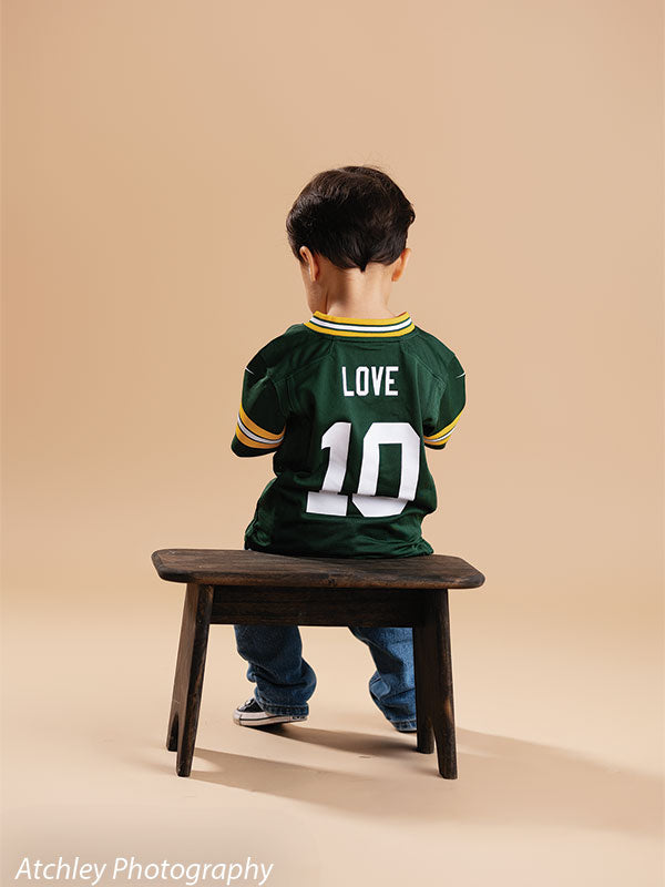 Toddler seated on a rustic wooden bench facing away from the camera, wearing a green football jersey with the name Love and number 10 on the back, blue jeans, and sneakers, photographed against a khaki brown seamless paper backdrop.