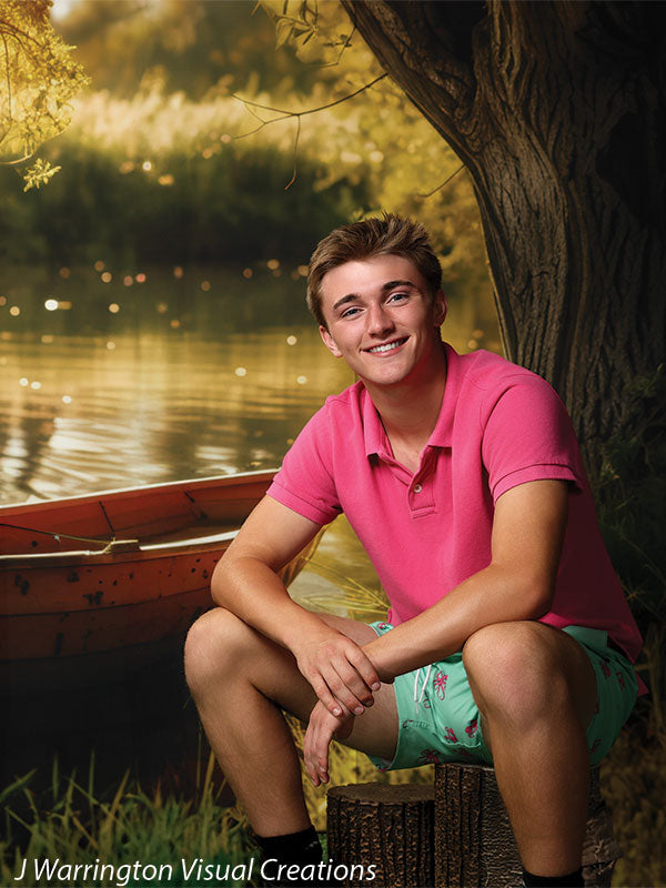 Teen boy in pink polo shirt smiling while seated in front of a scenic lakeside backdrop with a rowboat and tree, ideal for outdoor-themed studio portraits.