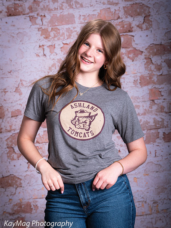 Teen girl in Ashland Tomcats t-shirt smiling confidently in front of a light pink distressed brick backdrop, perfect for casual senior or spirit portraits.