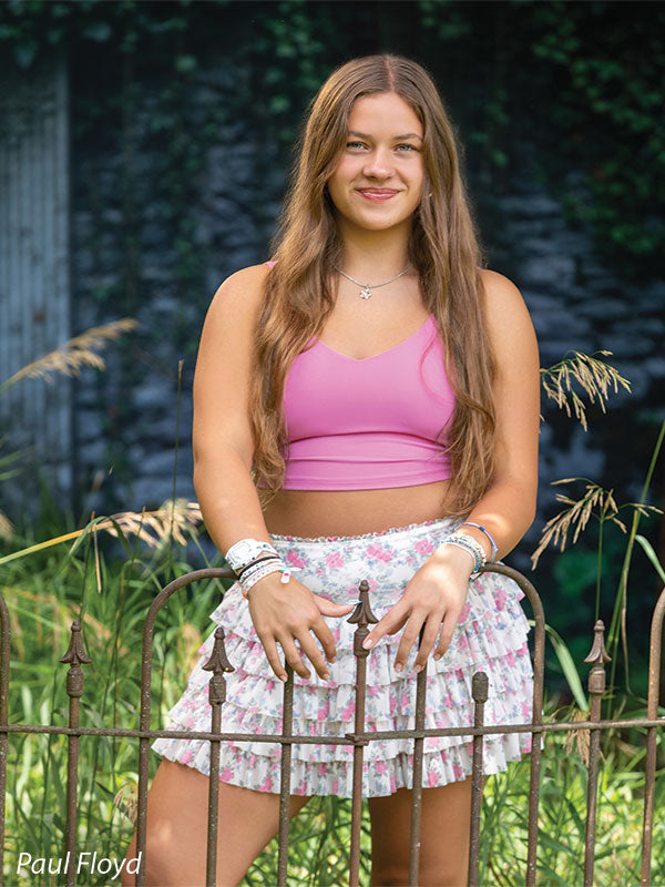 Lorian photography backdrop showing an ivy-covered classical garden wall with tall grasses and a wrought-iron fence, shown with a senior girl in a pink tank top and floral ruffle skirt resting her hands on the iron fence CP-8852