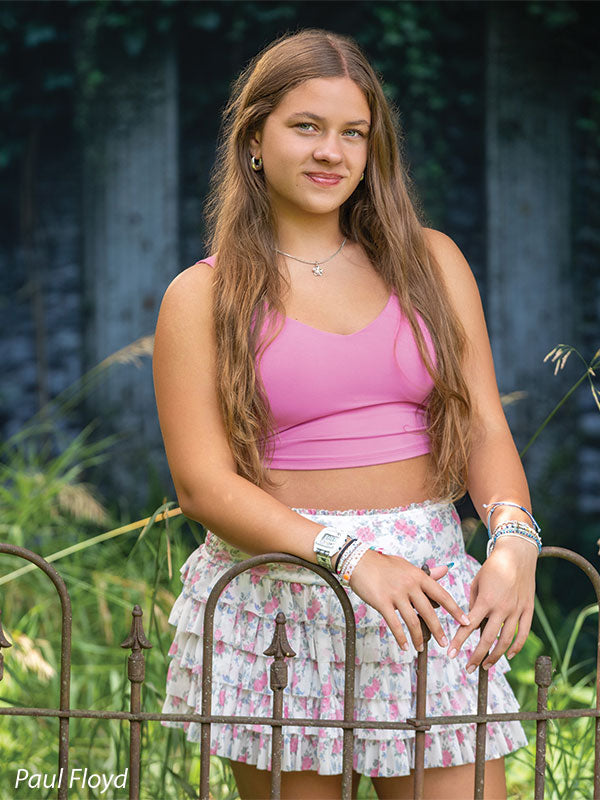 Lorian photography backdrop showing an ivy-covered classical garden wall with tall grasses and a wrought-iron fence, shown with a senior girl in a pink tank top and floral skirt turned slightly, smiling CP-8852