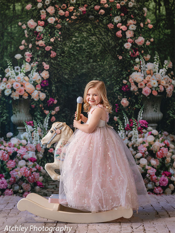 A young girl in a blush pink tulle dress smiles while sitting on a white rocking horse, posed against a lush rose garden backdrop with a floral arch and dense greenery.
