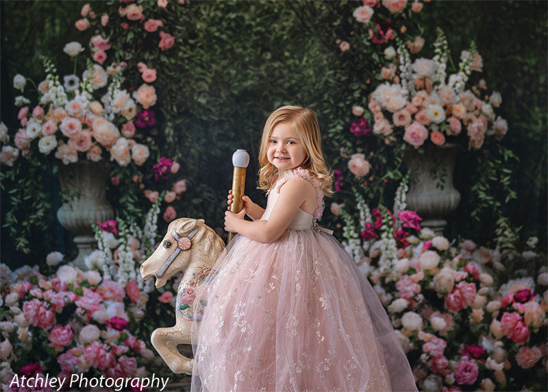 A young girl in a soft pink tulle dress smiles over her shoulder while seated on a white rocking horse, set against a dense rose garden backdrop with floral arrangements and greenery.
