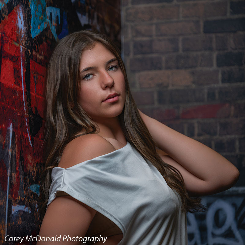 Marlowe Brick Wall Graffiti, photography backdrop panel for Denny StudioEdge silicone edge frame, shown with a senior girl in an off-shoulder white top posed against the graffiti wall SECP-8863