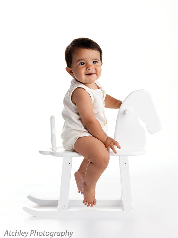 Smiling baby in a cream sleeveless romper sitting upright on a white wooden rocking horse in front of a minimalist white studio background, looking toward the camera with feet gently lifted and hands resting on the horse.
