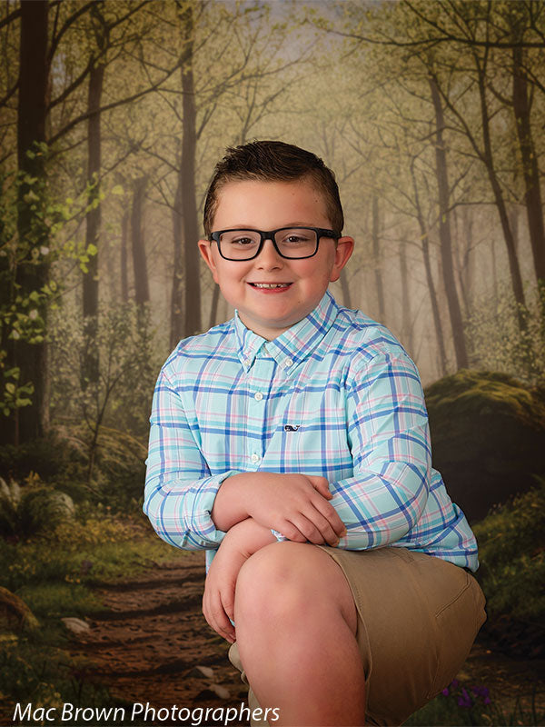 Young boy with short brown hair and glasses wearing a light blue plaid button-down shirt and khaki shorts, smiling while posed in front of a misty spring woodland forest photography backdrop featuring tall trees, soft morning haze, mossy rocks, and a winding dirt path.