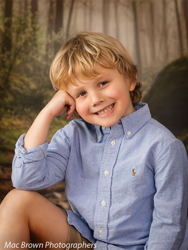Close-up portrait of a smiling young boy with blonde hair wearing a light blue button-down shirt, posed against a misty woodland spring portrait backdrop featuring soft-focus trees, greenery, and warm natural forest tones.