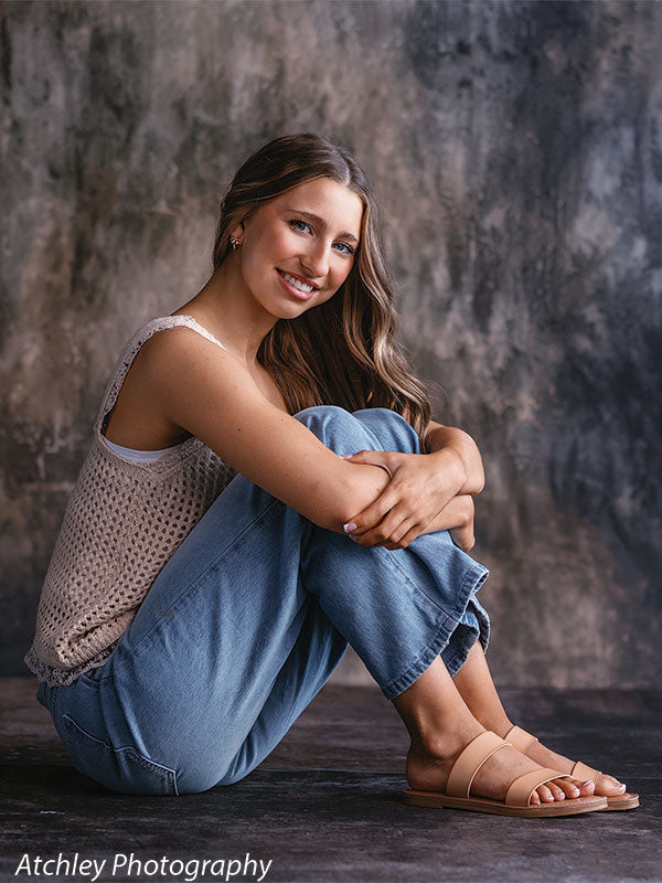Young woman seated on the floor with her knees pulled close, wearing a cream knit tank top, light blue jeans, and tan sandals, smiling toward the camera against a moody abstract brown earth tone backdrop with textured gray and warm brown variations.