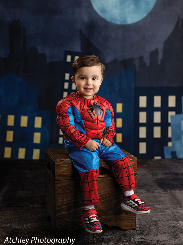 A smiling toddler wearing a red and blue superhero costume sits on a wooden box in front of a dark city skyline backdrop with glowing windows and a large moon overhead.
