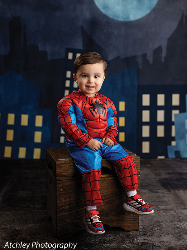 A toddler wearing a red and blue superhero costume sits on a wooden box, smiling, posed in front of a nighttime city skyline backdrop with glowing windows and a large moon.
