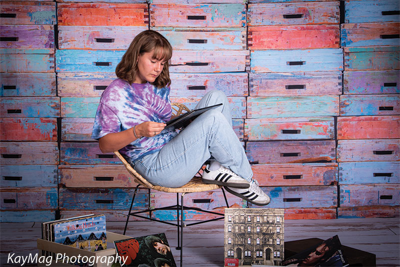 Teen girl in a tie-dye shirt examining vinyl records while seated in front of a multicolor wooden crate backdrop, styled for creative senior photography with a nostalgic vibe.