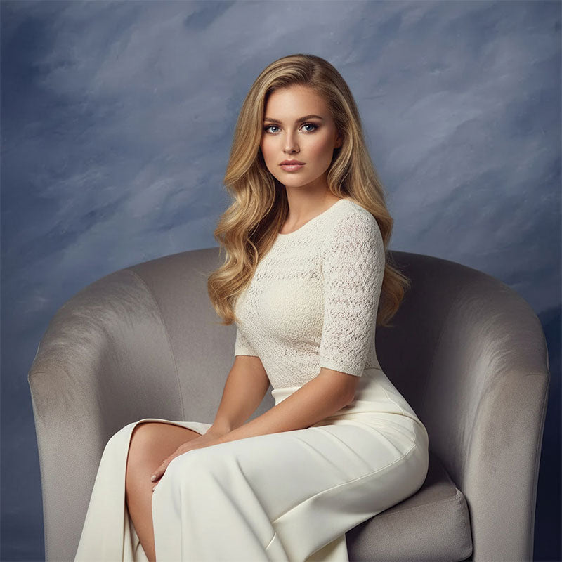 Blonde woman in a white lace top and cream skirt sits with hands resting on her lap on a curved gray chair in front of a muted blue gray mottled backdrop.