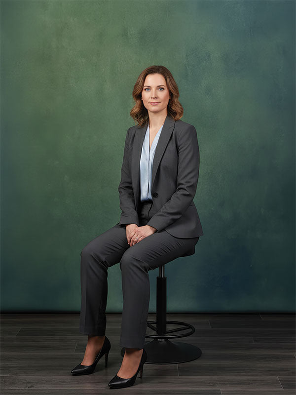 A woman in a gray suit sits upright on a stool, posed against a muted green professional studio backdrop with subtle texture and soft color depth.
