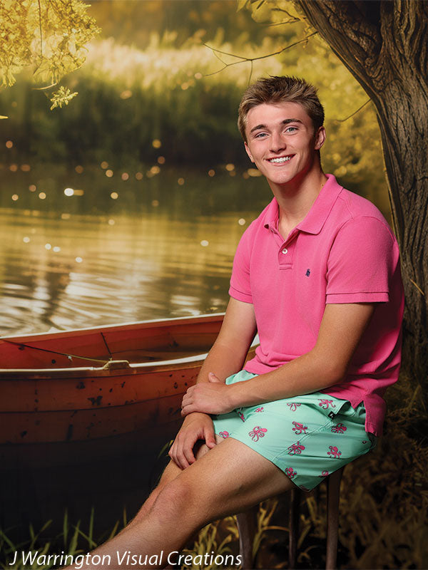 Teen boy seated near an orange rowboat with a peaceful pond and tree-lined backdrop, great for nature-themed senior photos or editorial shoots.