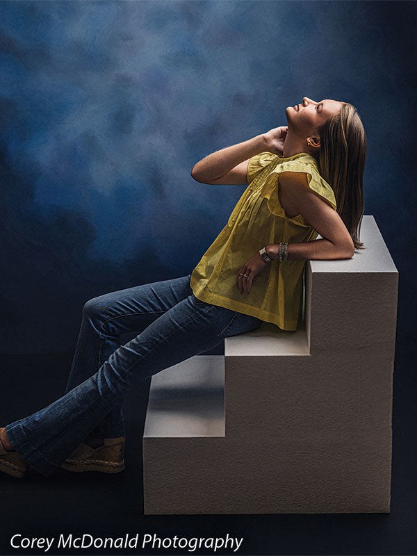 Neo Classical blue mottled texture photography backdrop shown with a senior girl in a yellow ruffle-sleeve top and blue flared jeans reclining against the white foam steps with a hand behind her head on white foam posing steps OM-1500